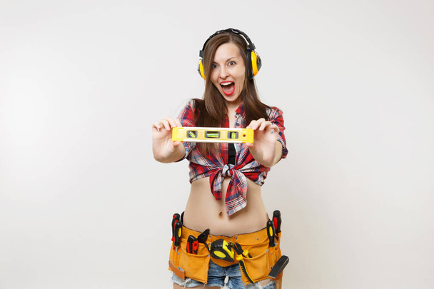 Pretty handyman woman in plaid shirt, denim shorts, noise insulated headphones, kit tools belt full of instruments hold building bubble spirit level isolated on white background. Female in male work - Photo, Image