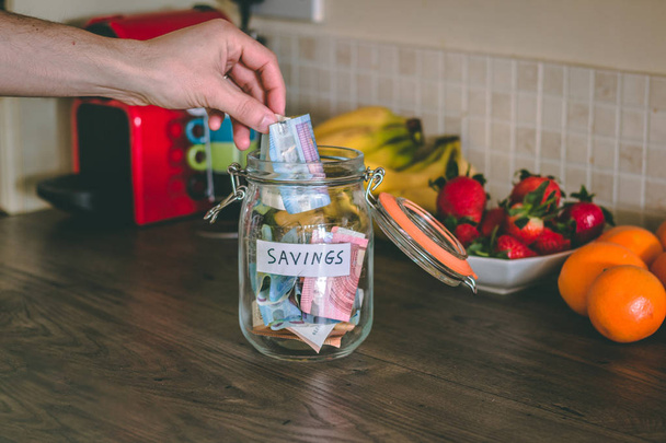 Concept: hand placing money on a savings jar on top of kitchen's wooden table - Fotoğraf, Görsel