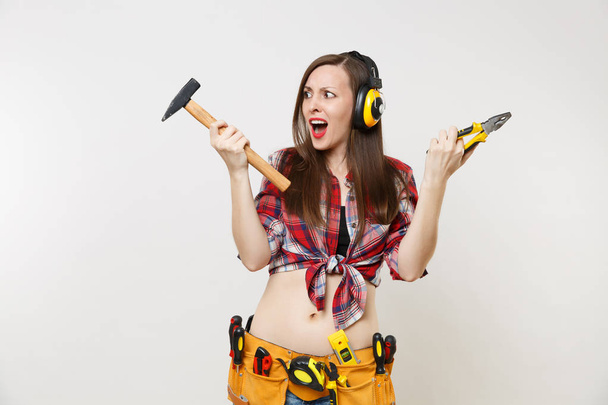 Angry emotional woman in tartan shirt, denim shorts, noise insulated headphones, kit tools belt full of instruments isolated on white background. Female in male work. Renovation occupation concept - Valokuva, kuva