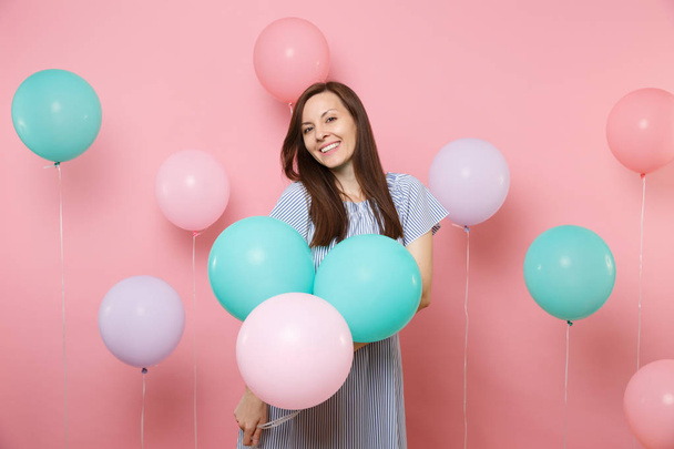Portrait of pretty smiling young happy brunette woman wearing blue dress holding colorful air balloons on bright trending pink background. Birthday holiday party, people sincere emotions concept - Фото, изображение