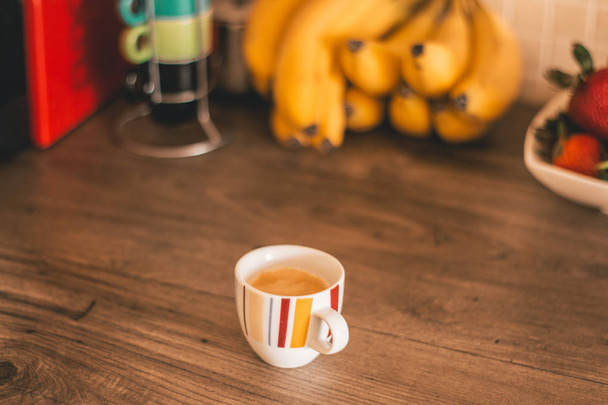 Full espresso cup on top of a wooden table with healthy fruit in the background - Foto, imagen