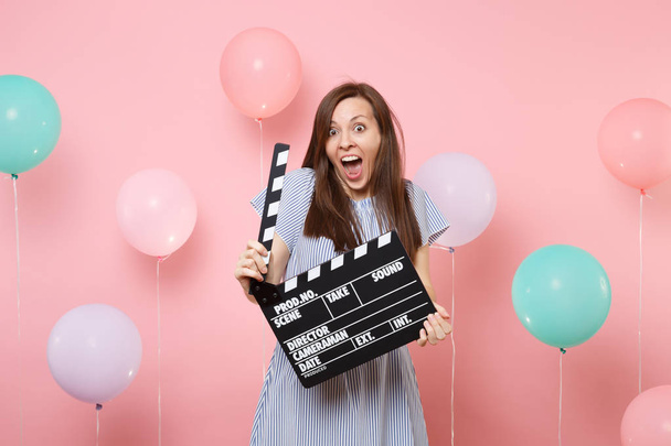 Портрет сумасшедшей возбужденной молодой женщины с открытым ртом в синем платье, держащей классический черный фильм, делающий доски на пастельно-розовом фоне с красочным воздушным шаром. Концепция празднования дня рождения
 - Фото, изображение