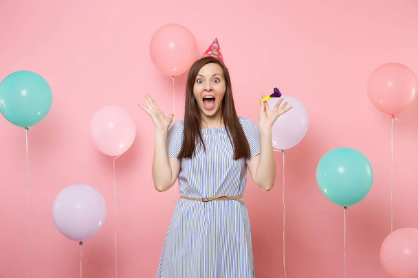 Portrait of surprised young woman with party whistle in birthday hat blue dress spreading hands screaming on pink background with colorful air baloons. Birthday holiday party, people sincere emotions - Photo, image