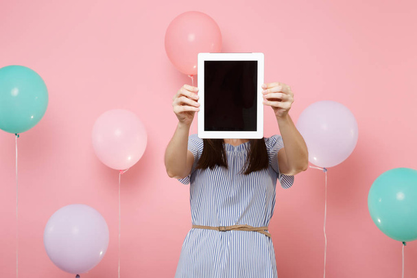 Portrait of young brunette woman wearing blue dress holding covering face with tablet pc computer with blank empty screen on pink background with colorful air balloons. Birthday holiday party concept - Φωτογραφία, εικόνα