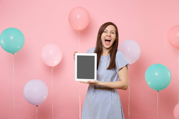 Portrait of beautiful overjoyed woman with opened mouth in blue dress holding tablet pc computer with blank empty screen on pink background with colorful air balloons. Birthday holiday party concept - Foto, Imagem