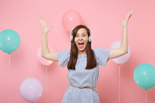 Portrait of overjoyed young woman with opened mouth with headphones wearing blue dress listening music spreading hands on pink background with colorful air balloons. Birthday holiday party concept - Photo, Image