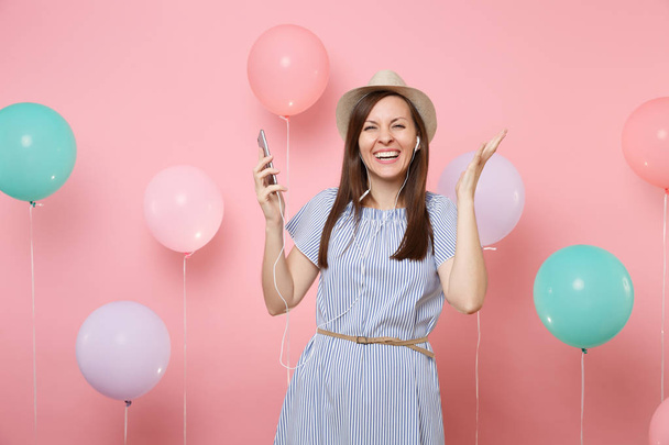 Portrét radostné mladé ženy v slaměné letní čepice a modré šaty s mobilní telefon a sluchátka poslouchat hudbu šíří ruku na růžovém pozadí s vrtulovým pohonem barevné. Oslava narozenin - Fotografie, Obrázek