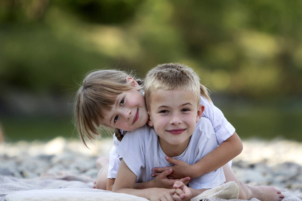 Двое молодых счастливых блондинок, улыбающихся детям, мальчик и девочка, брат и сестра лежали обнятые на галечном пляже на размытом ярком солнечном фоне летнего дня. Дружба и идеальный отдых
. - Фото, изображение