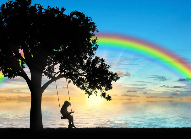 Silhouette of a little girl reading a book sitting on a swing by the sea with a rainbow. Conceptual image of childhood - Foto, afbeelding