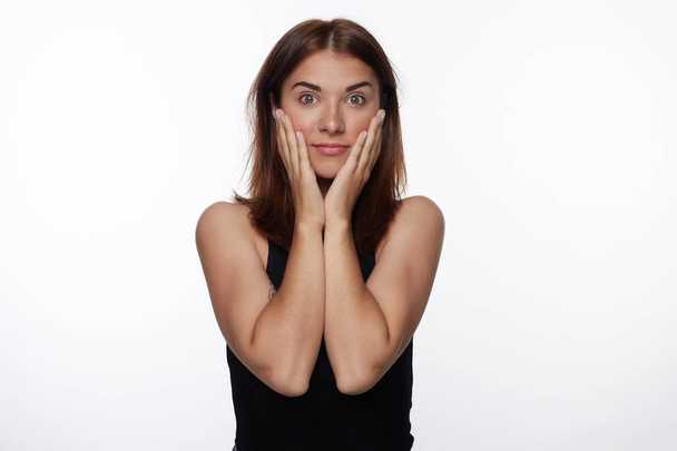 Human face expressions, emotions. Portrait of Caucasian female teenager with brown hair, wearing casual top, looking at the camera with surprised expression,holding hands on cheeks, smiling with eyes. - Photo, Image