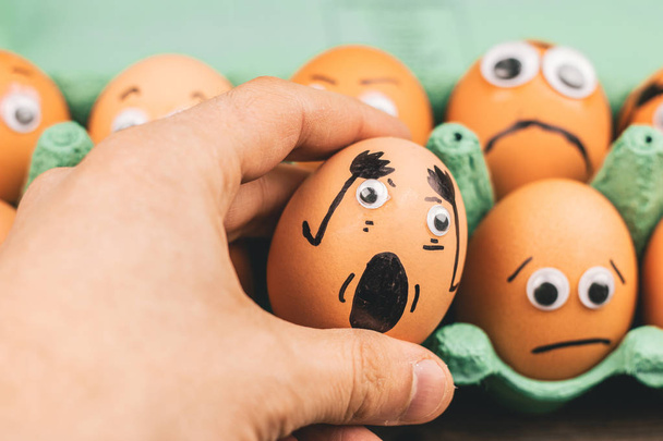 Fun concept: raw eggs with googly eyes and drawn features are in shock while sit in a green carton box and are being picked up by a hand - Fotografie, Obrázek