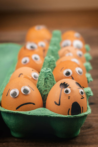 Concepto divertido: huevos crudos con ojos saltones y rasgos dibujados están en shock y tristes mientras se sientan en una caja de cartón verde encima de una mesa de madera
 - Foto, imagen