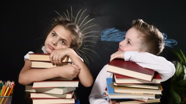 School concept. Cute boy and attractive girl sit with heaps of books. Schoolkids sit by desk. blackboard behind them. Schoolchildren hug books and feel sad - Footage, Video