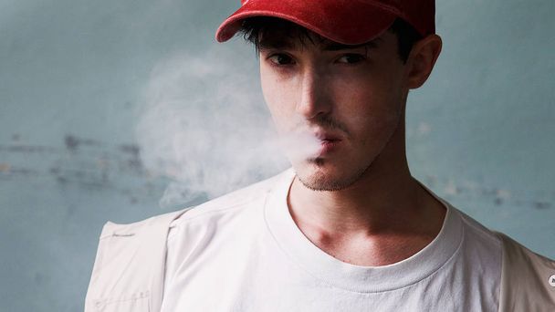 Retrato de un joven fumando con una gorra de béisbol roja y un impermeable. Retrato de moda de la modelo. Un hombre con una camisa blanca
. - Foto, imagen