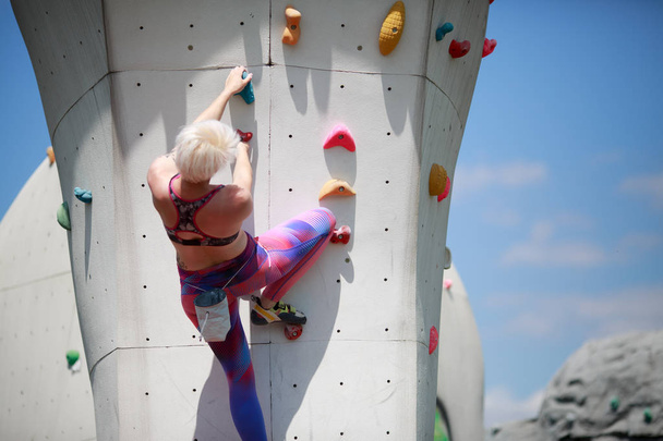 Photo from back of young girl with short haircut in sports clothes practicing on wall for rock climbing against blue sky with clouds in afternoon - Foto, Bild