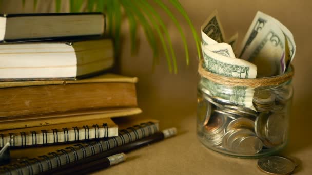 Boeken met glazen stuiver pot gevuld met munten en bankbiljetten. Collegegeld of onderwijs financiering concept. Beurs geld. - Video