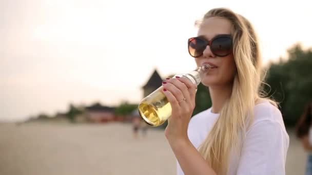 Young pretty woman walking and drinking beer of glass bottle on the beach during sunset, steadycam shot, slow motion. Blonde in glasses quenches thirst with lemonade beverage at sandy sea shore. - Footage, Video