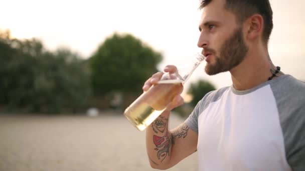 Young handsome tatooed bearded caucasian man drinking beer of glass bottle on the beach during sunset, steadycam shot, slow motion. Male quenches thirst with lemonade beverage at sandy sea shore. - Felvétel, videó