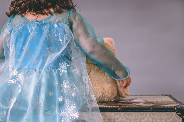 Une petite fille habillée en princesse tient un ours en peluche assis sur une poitrine et faisant face à un fond gris sans couture
 - Photo, image