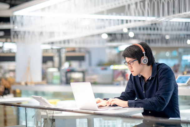 Jonge Aziatische zakenman muziek beluisteren door de koptelefoon tijdens het werken met de laptopcomputer in co-werkplaats. freelance of Digitaal Nomade levensstijl in stedelijke werkruimte concepten - Foto, afbeelding