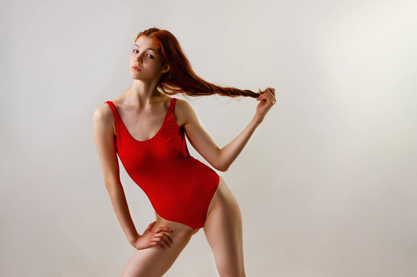 Jovem mulher magra sexy em maiô vermelho posando em estúdio. Retrato de moda de comprimento total de menina bonita com cabelo vermelho ondulado longo. Roupa de banho ou modo biquíni. Espaço de cópia, fundo cinza
 - Foto, Imagem
