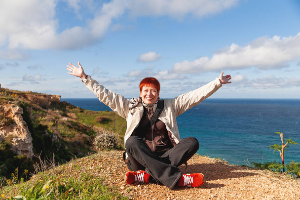 Happy wide smiling redhead women is sitting on the rock upon the beach. Gozo island, Malta. - 写真・画像