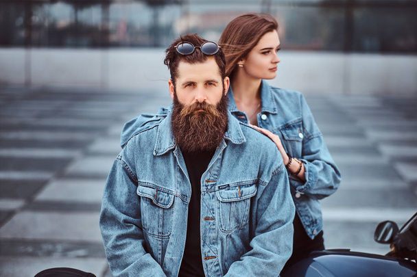 Gros plan portrait d'un couple hipster d'un homme barbu brutal et sa petite amie vêtus de vestes en jeans contre un gratte-ciel
. - Photo, image