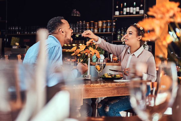Beautiful African-American couple in love having a great time together at their dating, an attractive couple enjoying each other, young woman feeding her man in a restaurant - 写真・画像