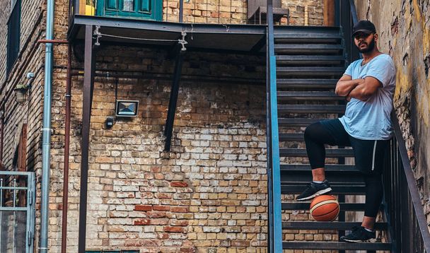 African-American bearded streetball player in cap dressed in a sportswear holds a basketball standing with crossed arms on stairs in a ghetto. - Fotografie, Obrázek
