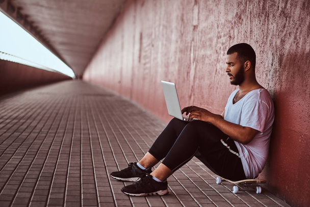 Portrait of an African-American bearded guy dressed in a white shirt and sports shorts working on a laptop while sitting on a skateboard under the bridge. - Φωτογραφία, εικόνα