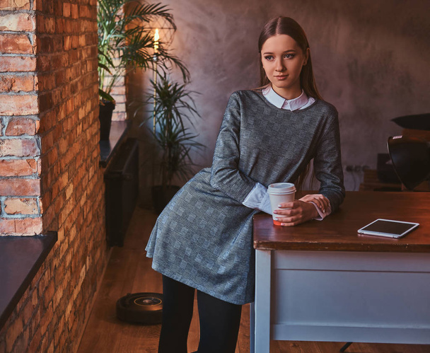 Chica joven vestida con un elegante vestido gris sostiene una taza de café para llevar mirando a una cámara mientras se apoya en la mesa en una habitación con interior loft
. - Foto, imagen