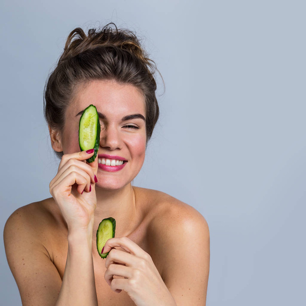 Smiling woman holding slices of cucumber in front of her eyes - Fotografie, Obrázek