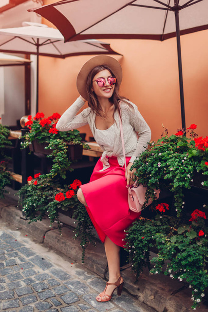 Молодая женщина в стильной одежде, украшениях и аксессуарах на открытом воздухе. Модель стоящая у кафе с цветами. Концепция красоты
 - Фото, изображение