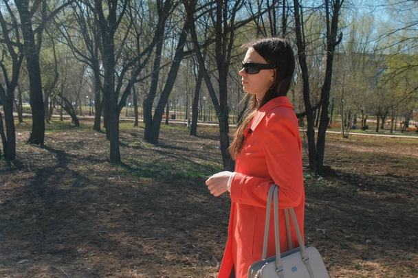 Young woman brunette in sunglasses in red coat walks in the spring Park. - 写真・画像