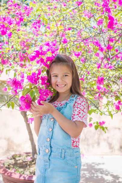 An adorable one little girl stands next to a large bush of pink flowers, bougainvillea - Fotoğraf, Görsel