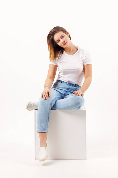 Girl teenager in jeans sits in cube on white studio, full body - Foto, imagen