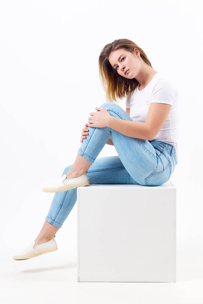 Girl teenager in blue jeans sits on cube in white studio, full body - Фото, изображение