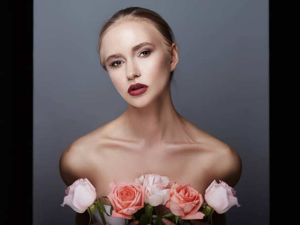 Blonde girl holding rose flowers near her face. Beauty portrait of a woman on a dark background. Perfect makeup, beautiful body. Clean smooth skin on the girl's face. Body cosmetics, skin care - Фото, изображение