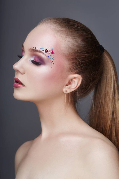 Art makeup over the eyebrows of women many rhinestones of different shapes, beautiful face smooth skin care. Beauty makeup on the woman face close-up. Professional makeup artist, long beautiful hair - Foto, Imagem