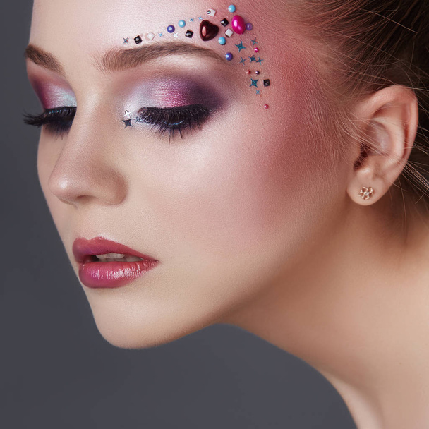 Art makeup over the eyebrows of women many rhinestones of different shapes, beautiful face smooth skin care. Beauty makeup on the woman face close-up. Professional makeup artist, long beautiful hair - Photo, image