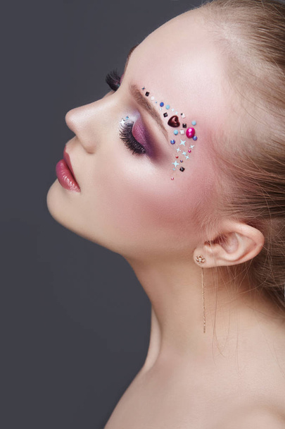 Art makeup over the eyebrows of women many rhinestones of different shapes, beautiful face smooth skin care. Beauty makeup on the woman face close-up. Professional makeup artist, long beautiful hair - Zdjęcie, obraz