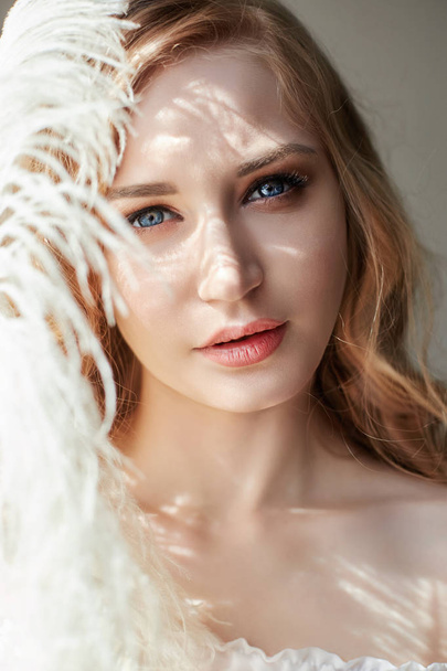 Girl in white light dress and curly hair with big feather near her face, portrait woman with feather, purity and innocence. Curly blonde looks romantic, beautiful eyes. White ostrich feather in hands - Fotografie, Obrázek