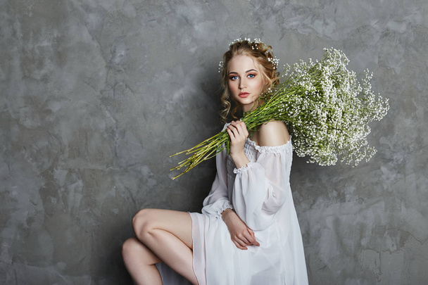Girl white light dress and curly hair, portrait of woman with flowers at home near the window, purity and innocence. Curly blonde romantic look, beautiful eyes. White wildflowers in hands - Foto, Imagem