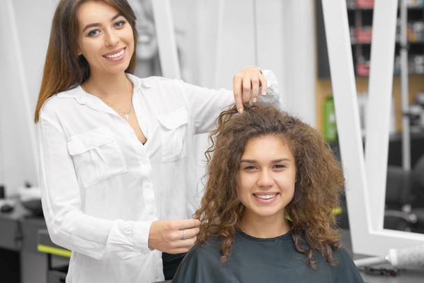 Smiling female hairstylist working with young curly client, looking at camera, laughing. Hairdresser wearing white casual shirt, girl is covered with special black cape. Posing in whitish beaty salon. - Zdjęcie, obraz