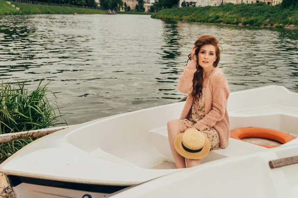 the girl is sitting in a white boat on the river. dressed in a flowered dress, a beige sweater, in hands holds a hat from the sun. summer vacation. - Foto, immagini