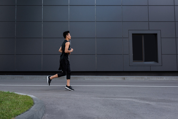 side view of young asian sportsman in earphones running at city street - 写真・画像
