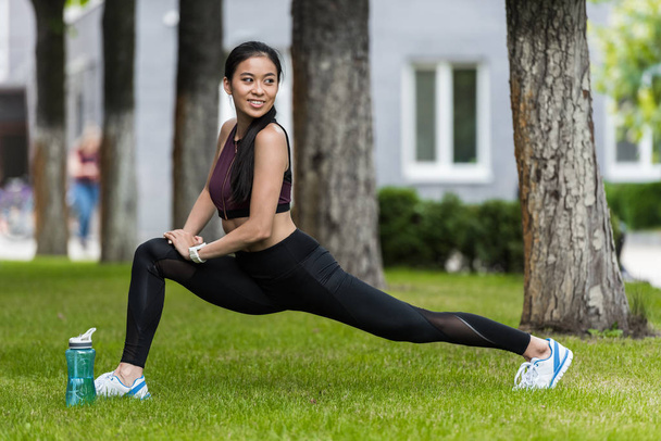 smiling asian female athlete stretching near sport bottle of water on grass in park  - Zdjęcie, obraz