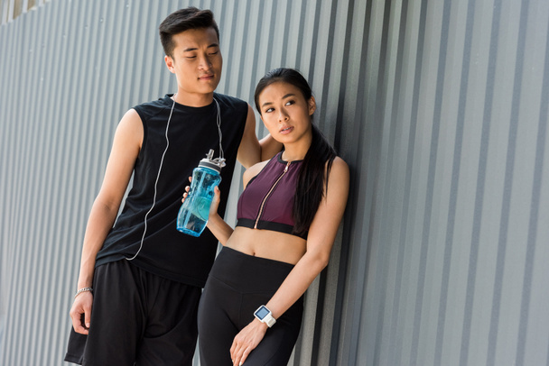 lächelnder junger asiatischer Sportler mit Kopfhörern über dem Hals, der neben Sportlerin mit Wasserflasche auf der Straße steht - Foto, Bild