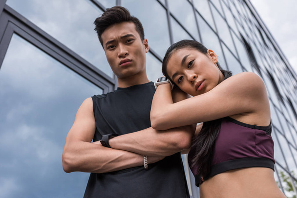 low angle view of serious asian sportsman with crossed arms and sportswoman standing together and looking at camera at urban street - Photo, Image