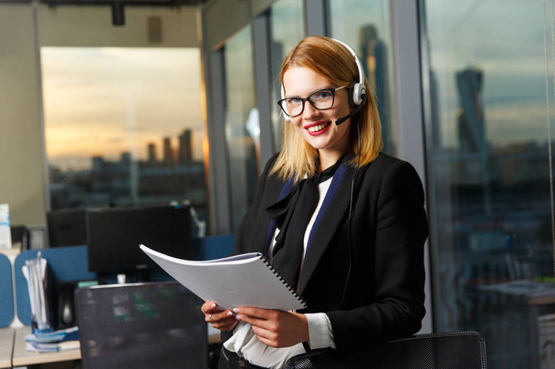 Photo of smiling woman with glasses and headphones with paper in hands near glass wall - Foto, Imagem
